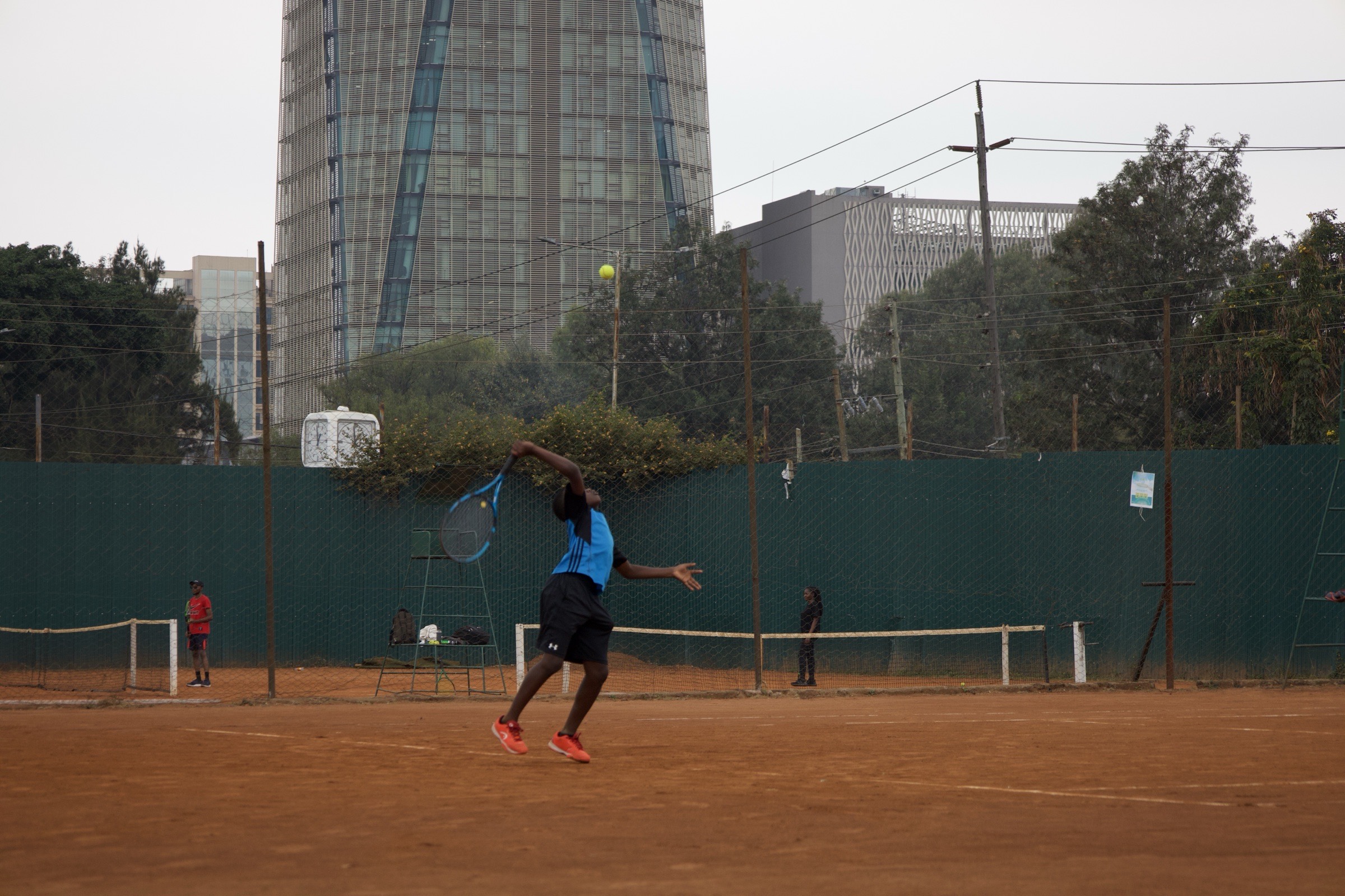 Young athlete mid-serve on a clay court with the Nairobi skyline behind them